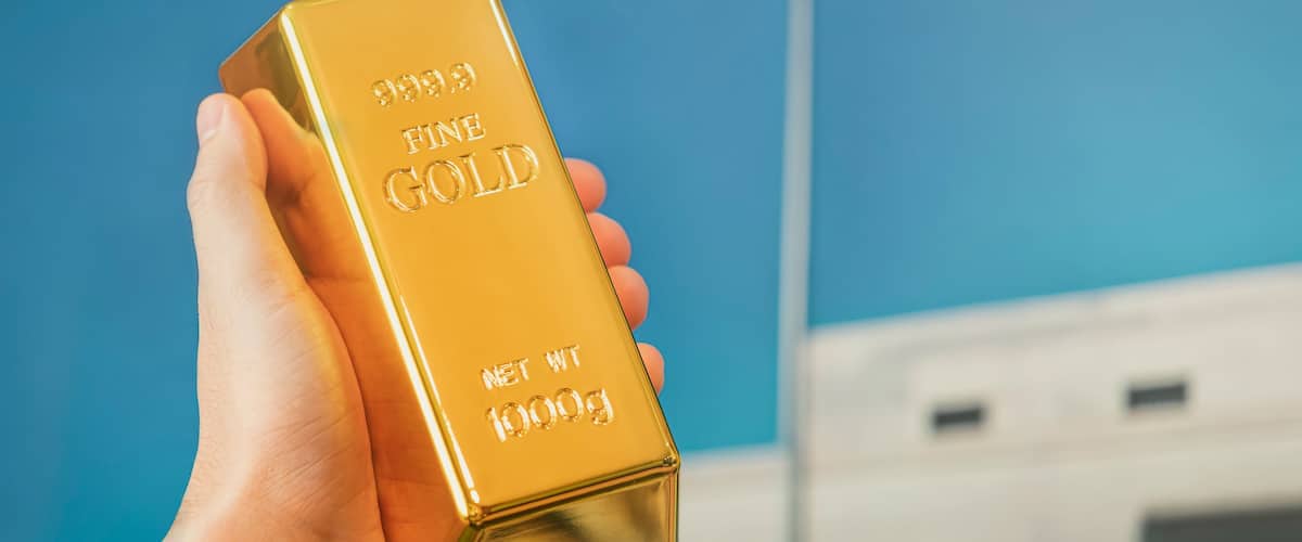 A hand holding a gold bar with "999.9 Fine Gold" and "NET WT 1000g" inscriptions.