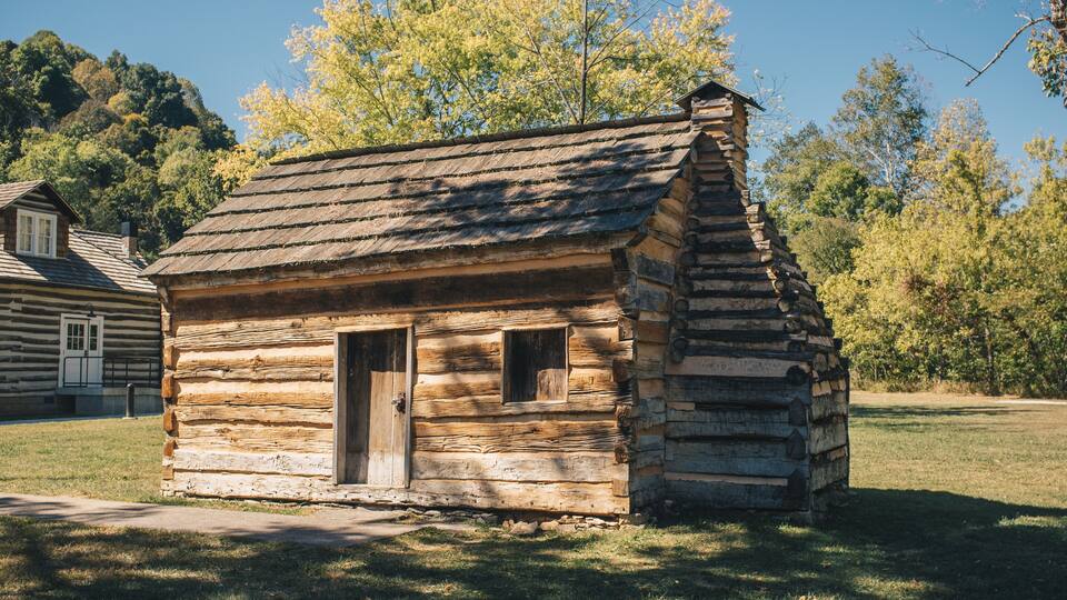 A small log cabin with a wooden door and windows, surrounded by grass and trees.