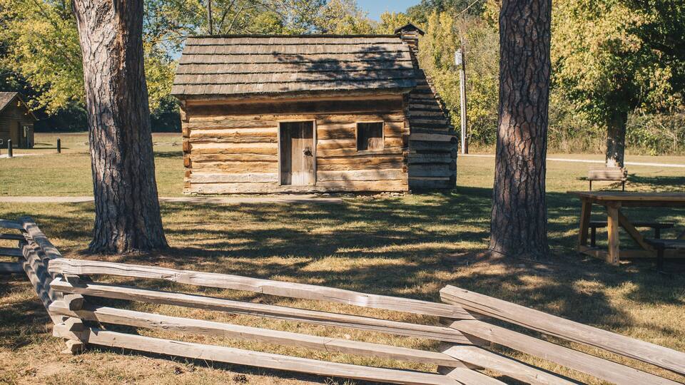 A rustic log cabin with a wooden door and window, surrounded by trees and a picnic table.