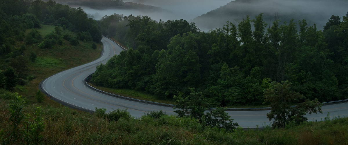 A winding road through a forested valley with mist-covered mountains in the background.
