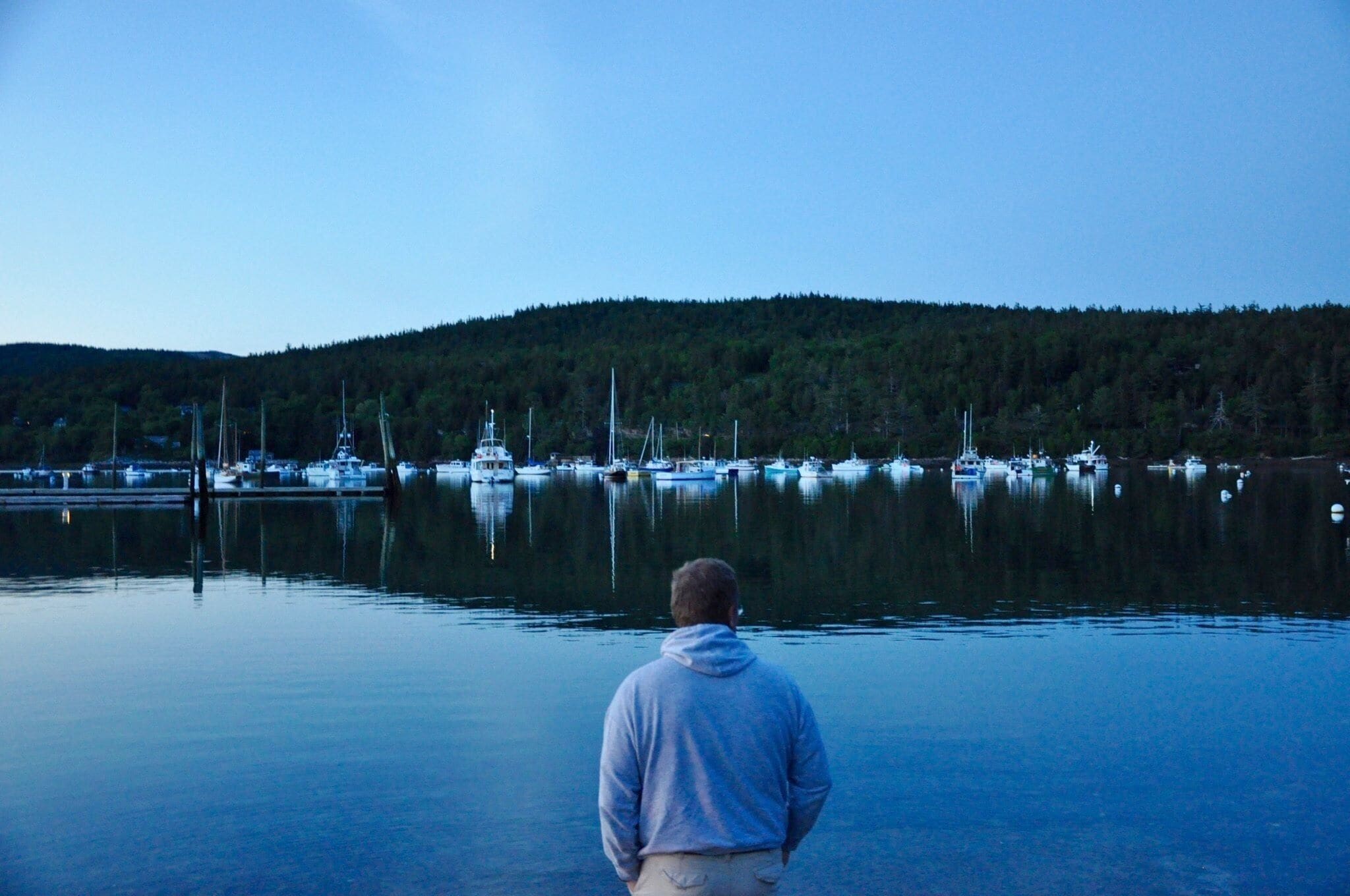 A man standing by a calm lake with several boats anchored in the distance and a forested hill in the background.