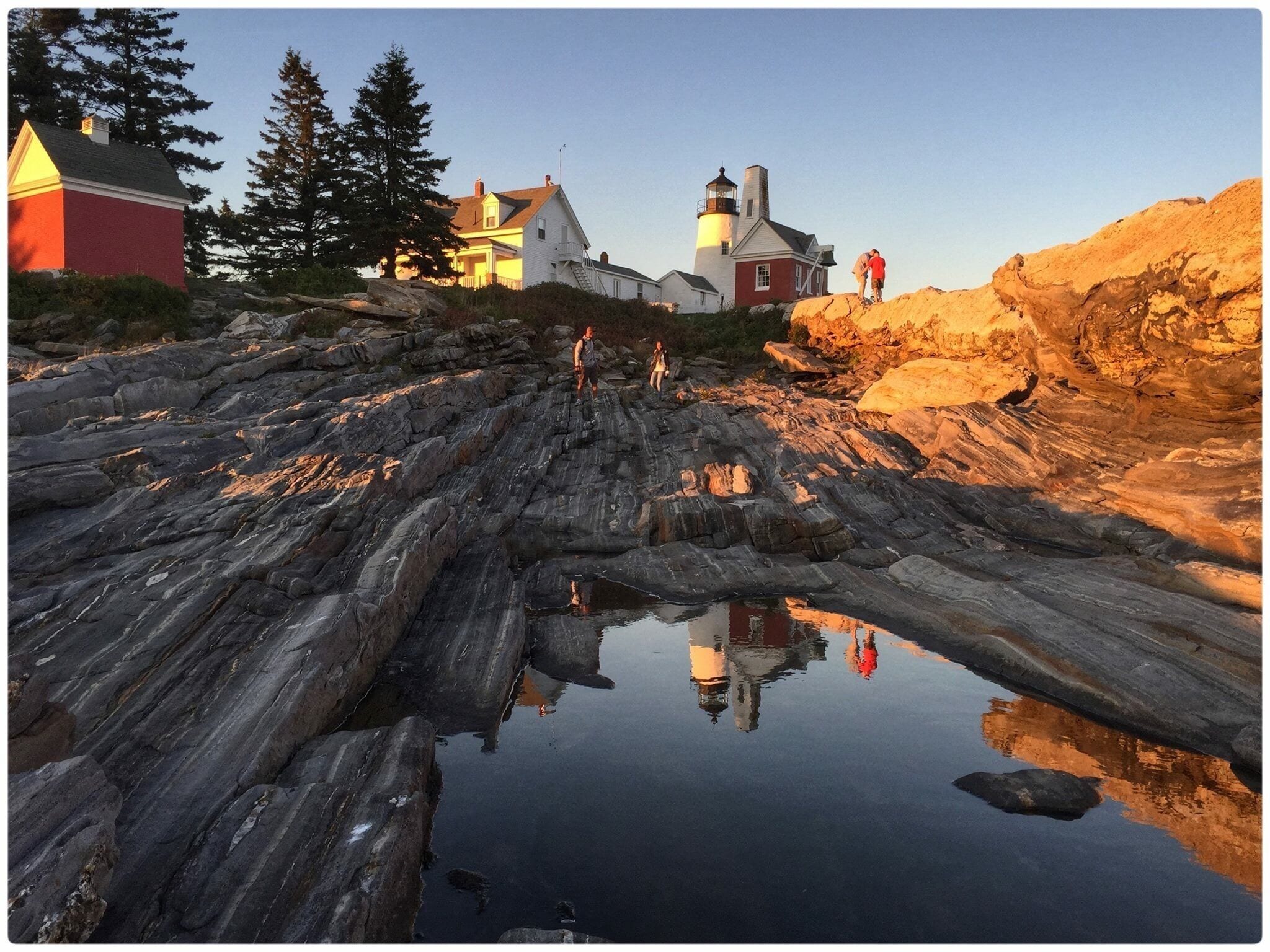 A rocky shoreline with a lighthouse, a red house, and a person walking.