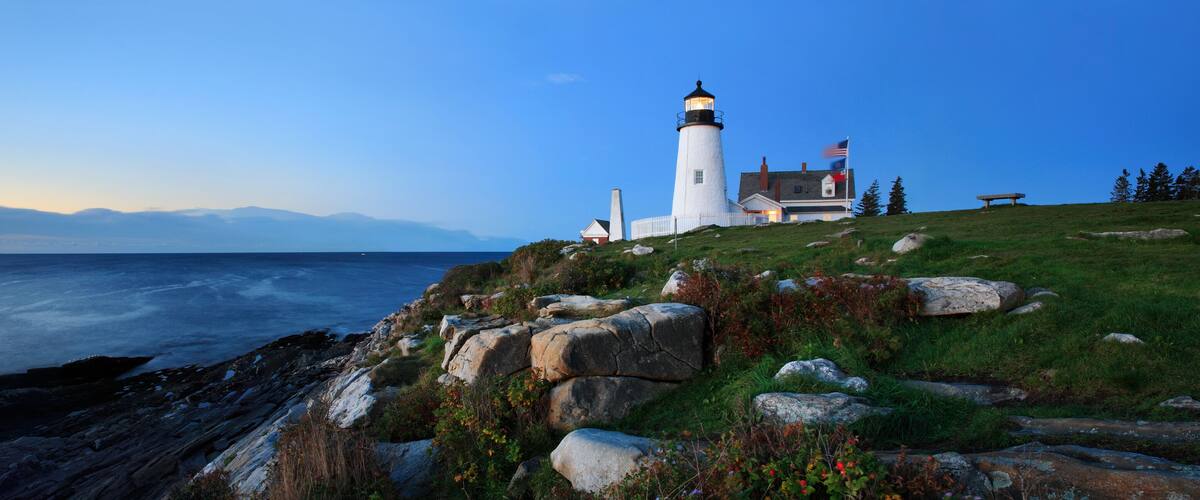 A lighthouse on a grassy hill overlooking the ocean with mountains in the background.