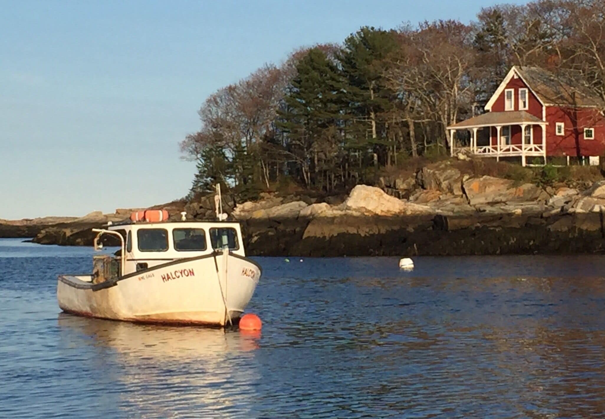 A boat named HALCYON anchored near a red house with a white porch, surrounded by trees and rocks.