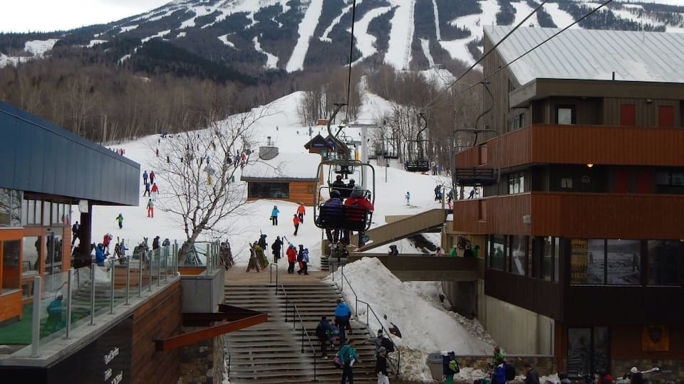 A ski lift at a mountain resort with a snowy landscape and buildings in the foreground.