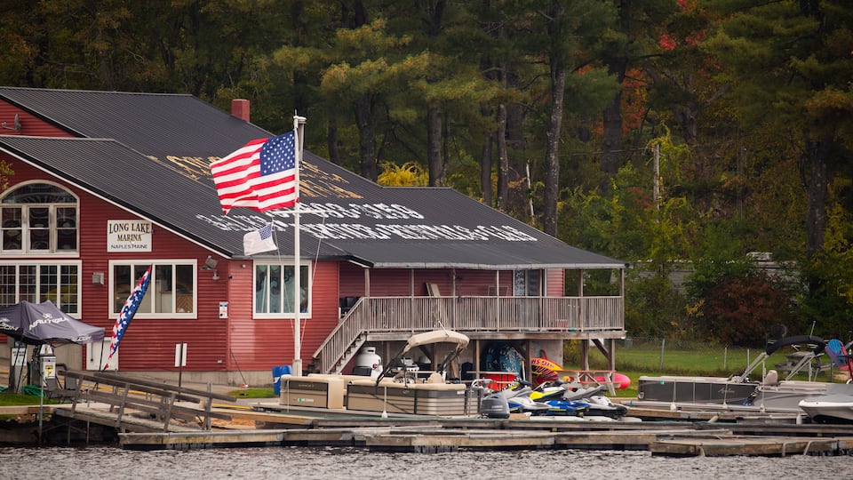 A red building with an American flag, a dock with boats, and a forest in the background.