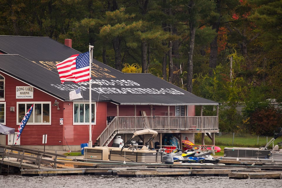 A red building with an American flag, a dock with boats, and a forest in the background.