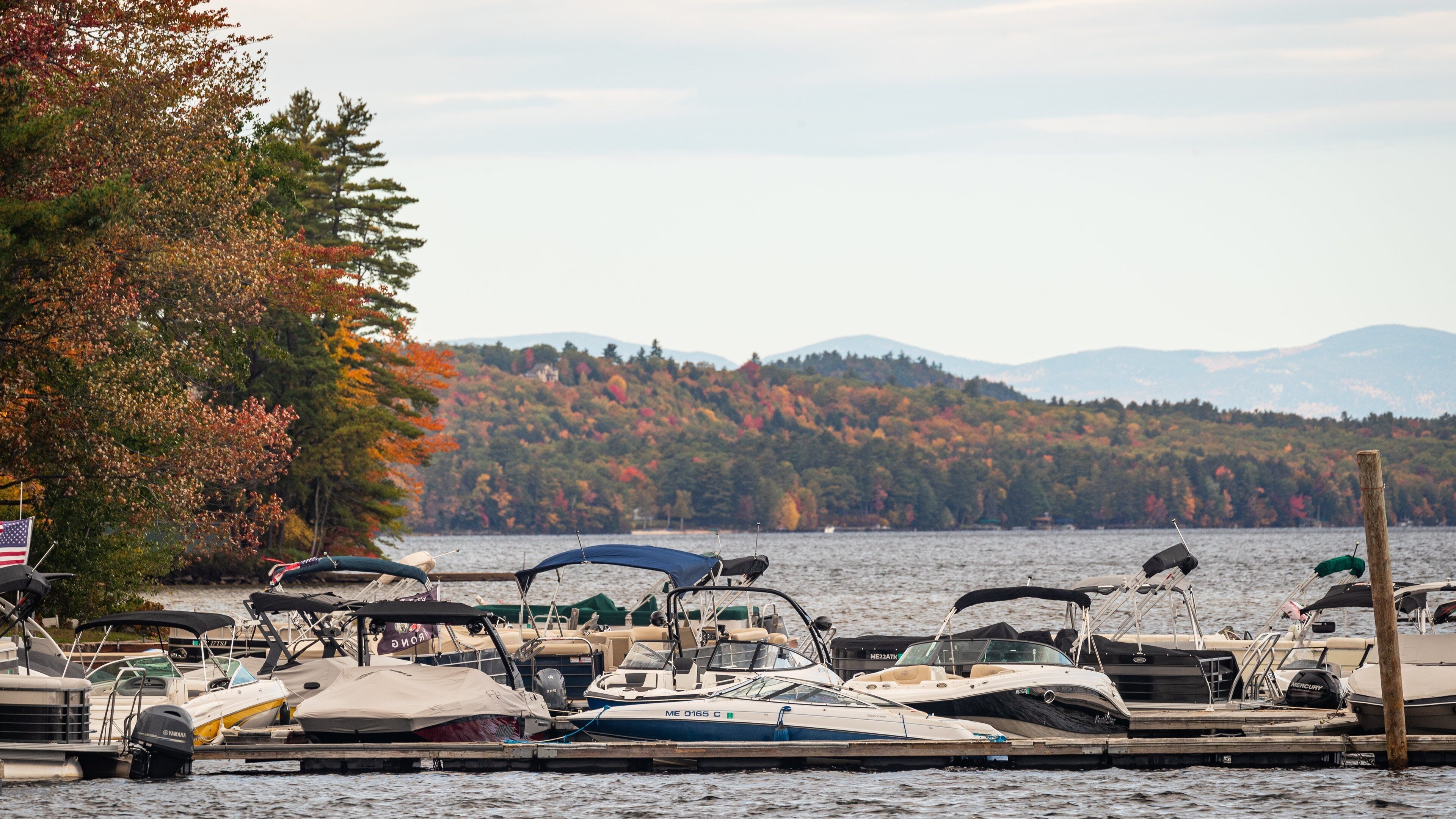 A marina with boats docked, surrounded by autumn-colored trees and a lake with mountains in the background.
