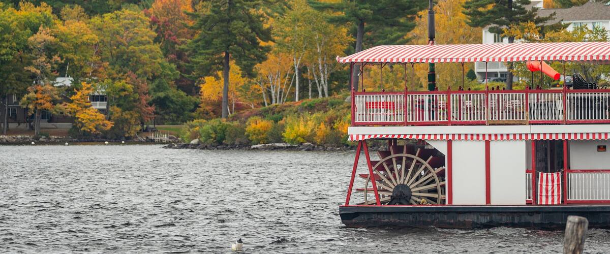 A houseboat with a red and white striped awning and a large water wheel on a lake surrounded by autumn-colored trees.