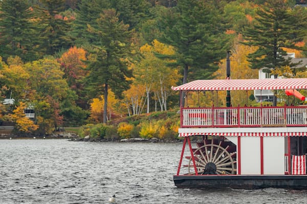 Une péniche dotée d’un auvent rayé de rouge et de blanc et d’une grande roue à aubes, située sur un lac bordé d’arbres aux couleurs automnales.
