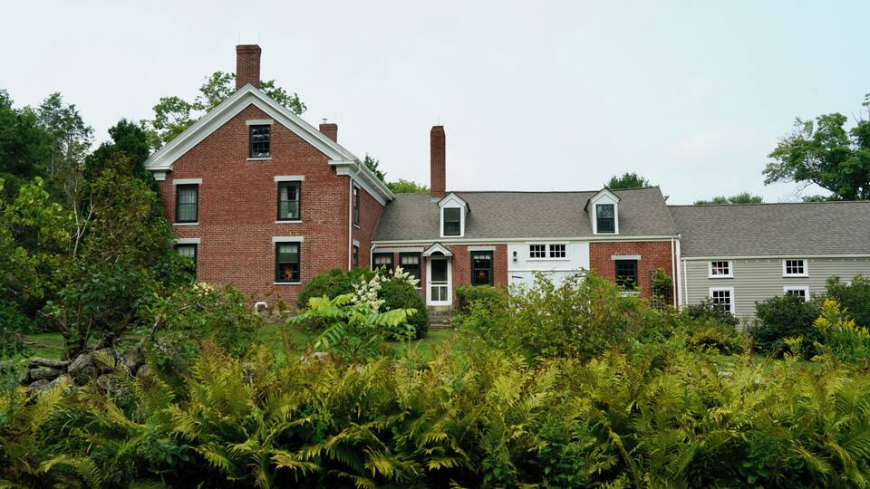 A large brick house with a gabled roof and a chimney, surrounded by lush greenery and trees.