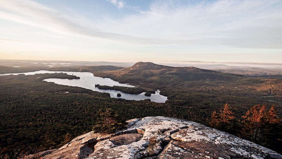 A mountainous landscape with a lake surrounded by dense forest and a prominent rocky outcrop in the foreground.