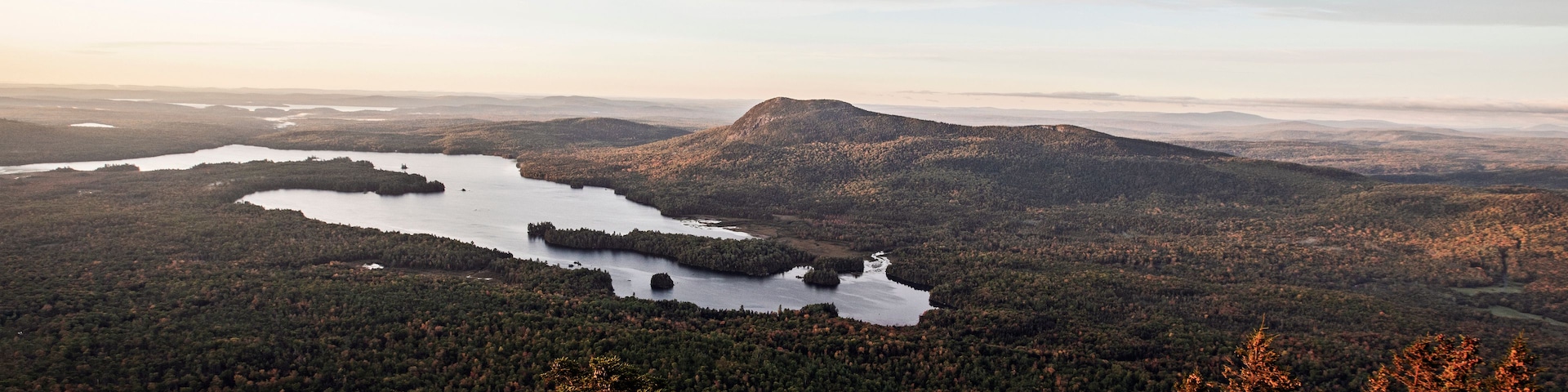 Paisagem montanhosa com um lago rodeado por floresta densa e um afloramento rochoso em primeiro plano.