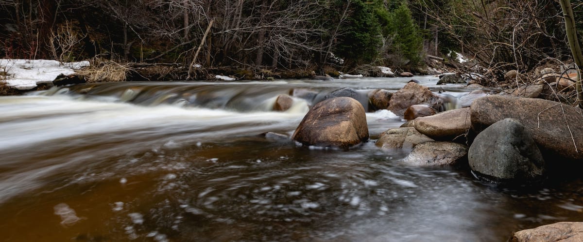 A flowing river with large rocks, surrounded by trees and a snowy bank.
