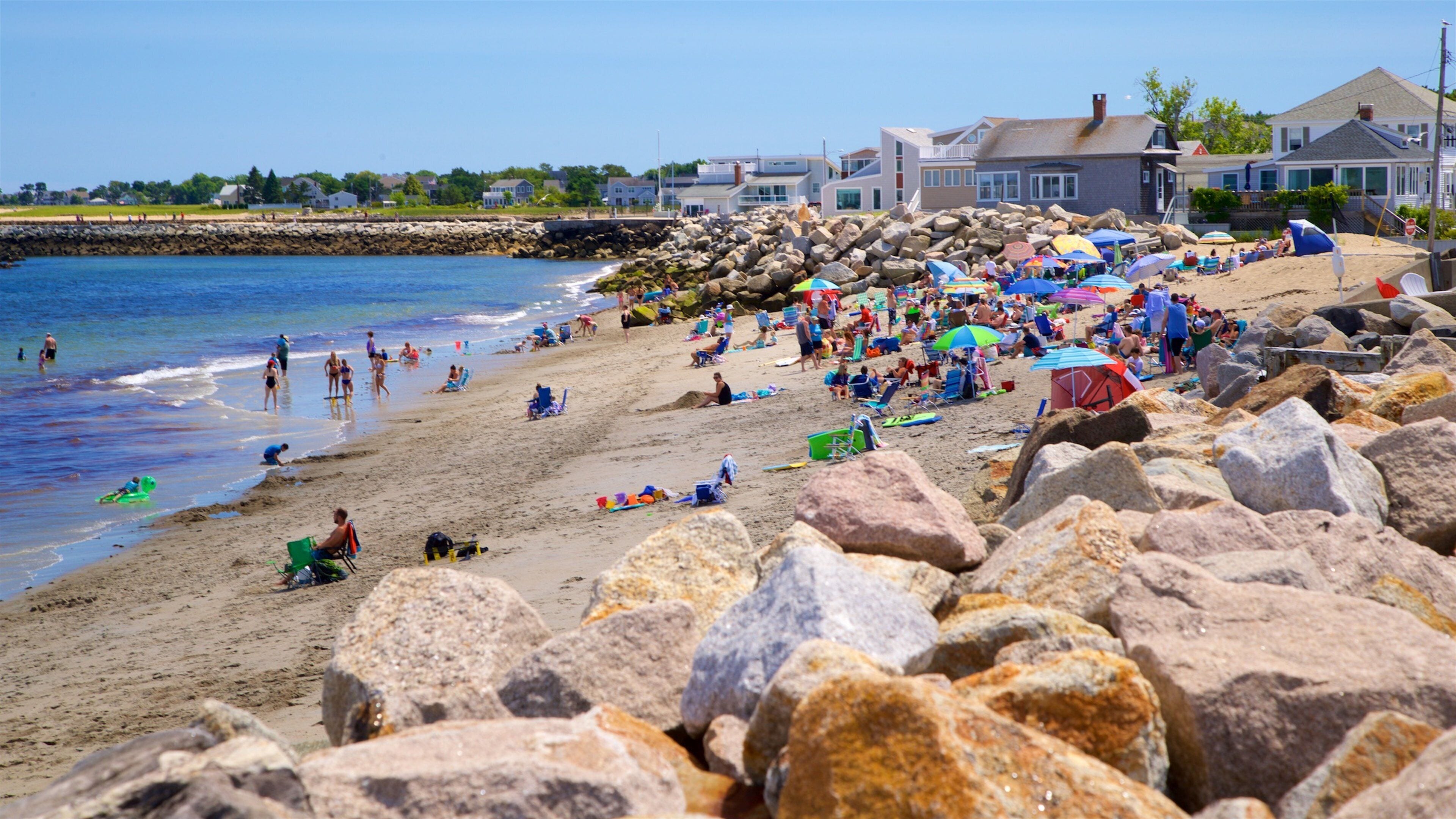 A crowded beach with people sunbathing and swimming, large rocks in the foreground, and houses in the background.