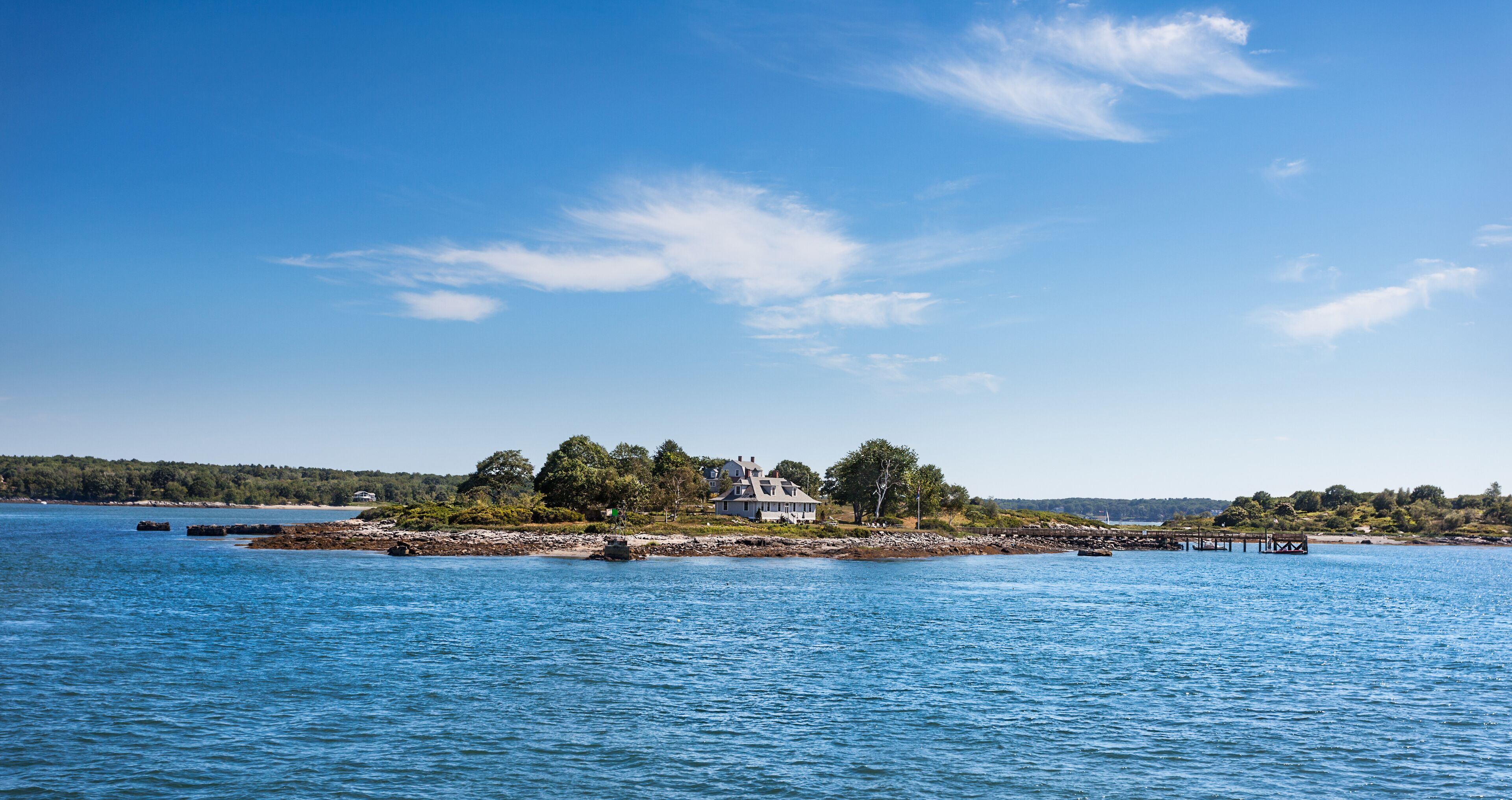A coastal landscape with a house surrounded by trees, a rocky shoreline, and calm blue waters.