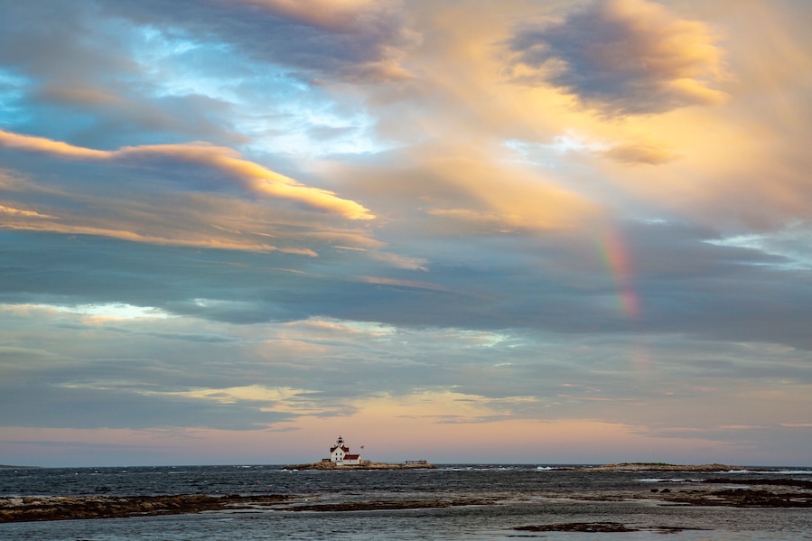 A lighthouse on a rocky outcrop with a rainbow in the sky.