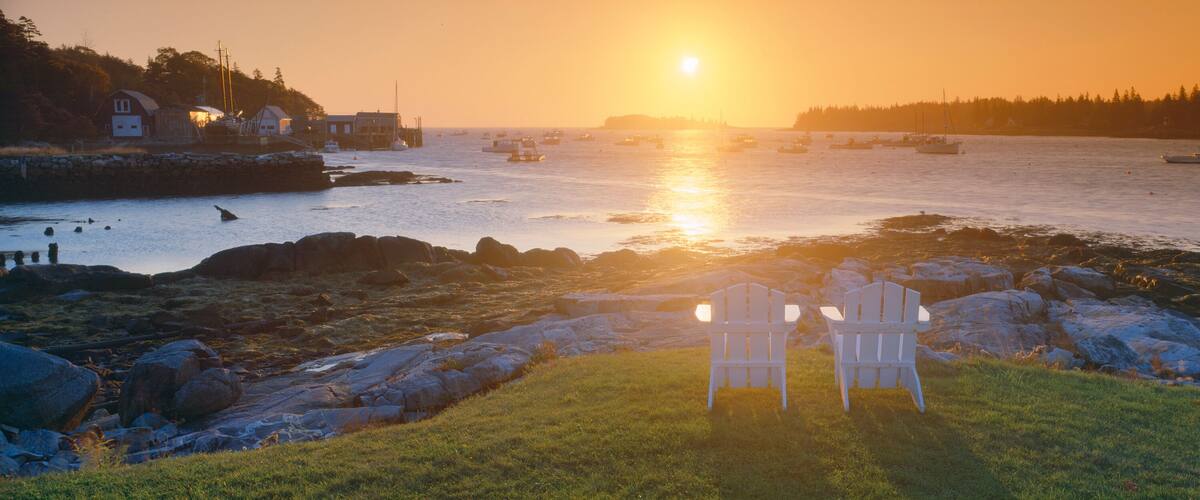 A rocky shoreline with two white chairs facing the ocean at sunset. Boats are visible on the water, and a forested hill is in the background.