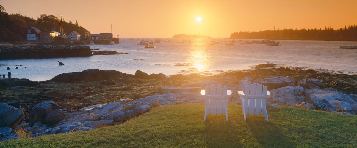 A sunset over a rocky shoreline with chairs facing the water, boats in the distance, and a forested hill.