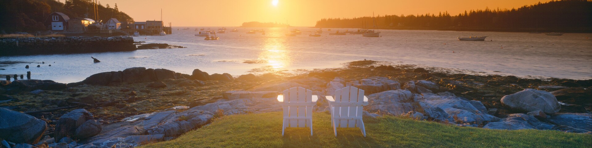 A sunset over a rocky shoreline with chairs facing the water, boats in the distance, and a forested hill.