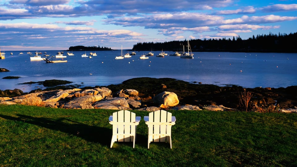 This is an image of two white lawn chairs facing toward the nearby harbor. There are fishing boats moored in the harbor in this small lobster village. The chairs are surrounded by green grass and the harbor has clear blue water. The sky is blue with white clouds.