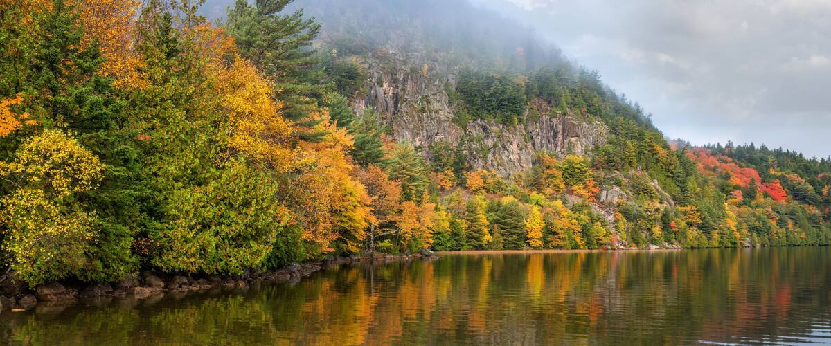 A calm lake surrounded by autumn-colored trees and a rocky mountain in the background.