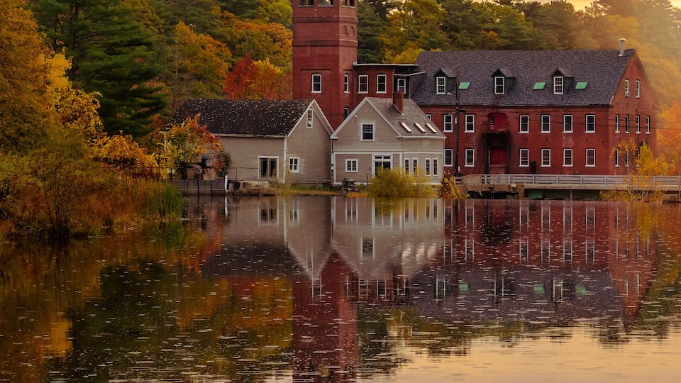 A historic building with a tower, surrounded by autumn trees and reflecting in a calm lake.