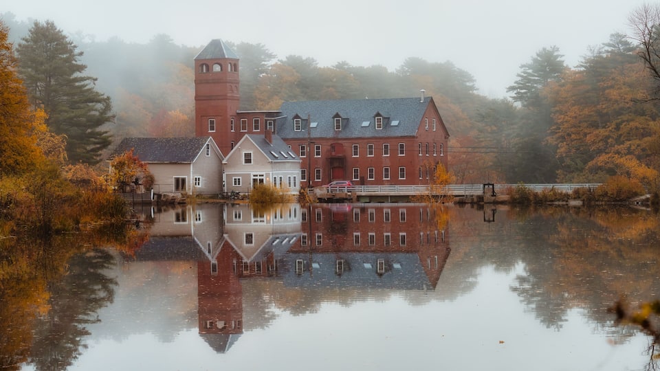 A historic building complex with a prominent tower, reflecting in a calm lake surrounded by autumn foliage.