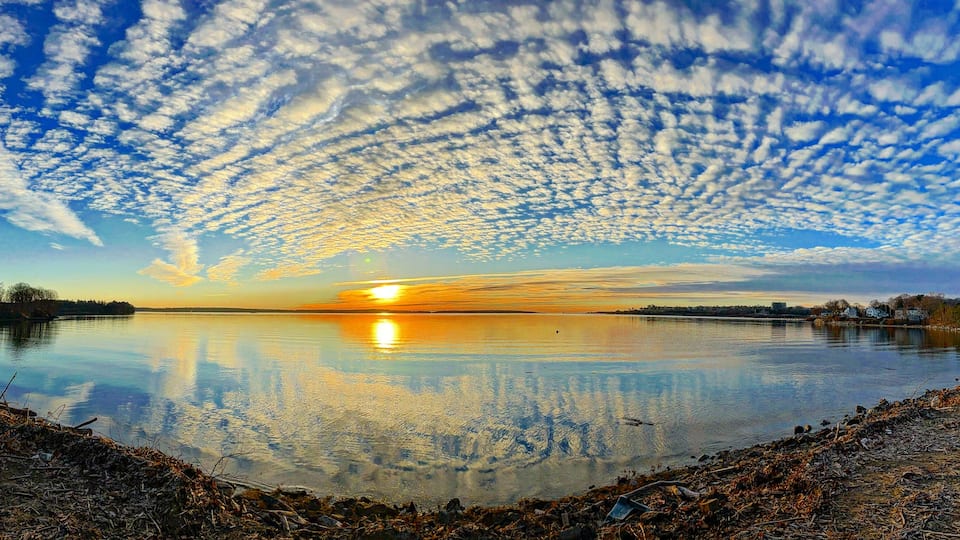 A sunset over a calm lake with a reflection of the sky and clouds, surrounded by a shoreline with sparse vegetation and a distant shoreline with buildings.
