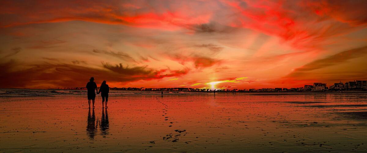A beach with a sunset, silhouettes of people, and houses in the background.