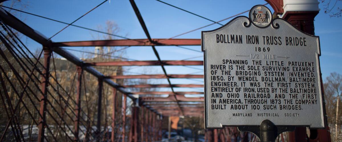 A historical sign for the Bollman Iron Truss Bridge, built in 1869, with details about its significance and construction.