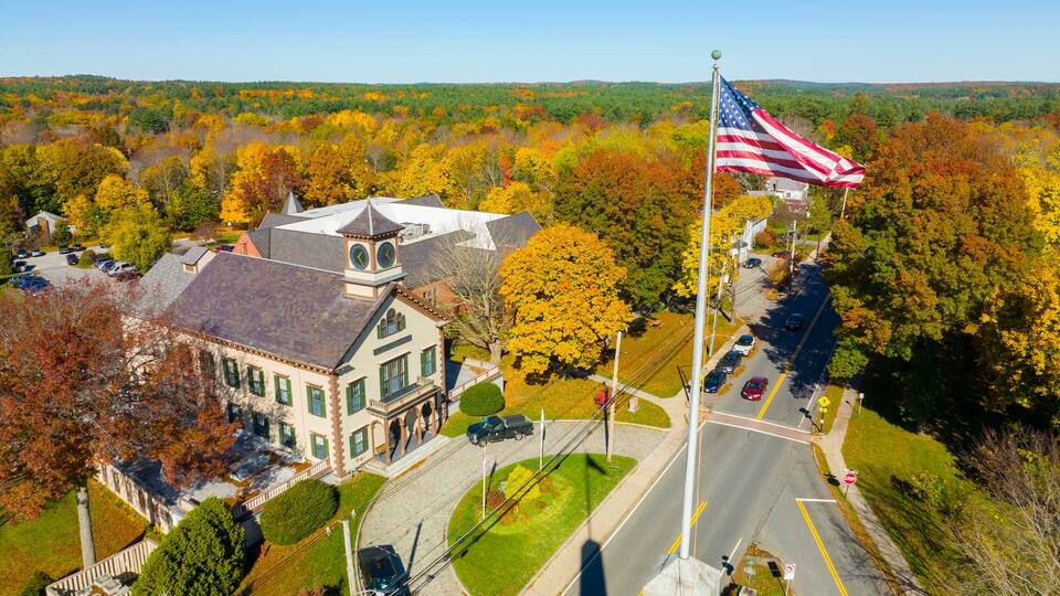An aerial view of a town with a prominent building, a flagpole with an American flag, and autumn-colored trees.