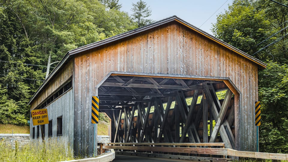 A wooden covered bridge with a 'DANGER KEEP OFF DAM' sign, surrounded by lush greenery.