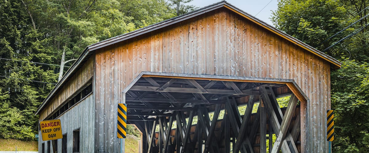 A wooden covered bridge with a 'DANGER KEEP OFF DAM' sign, surrounded by lush greenery.