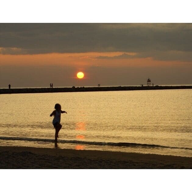 Un atardecer sobre una playa con un niño jugando en el agua.