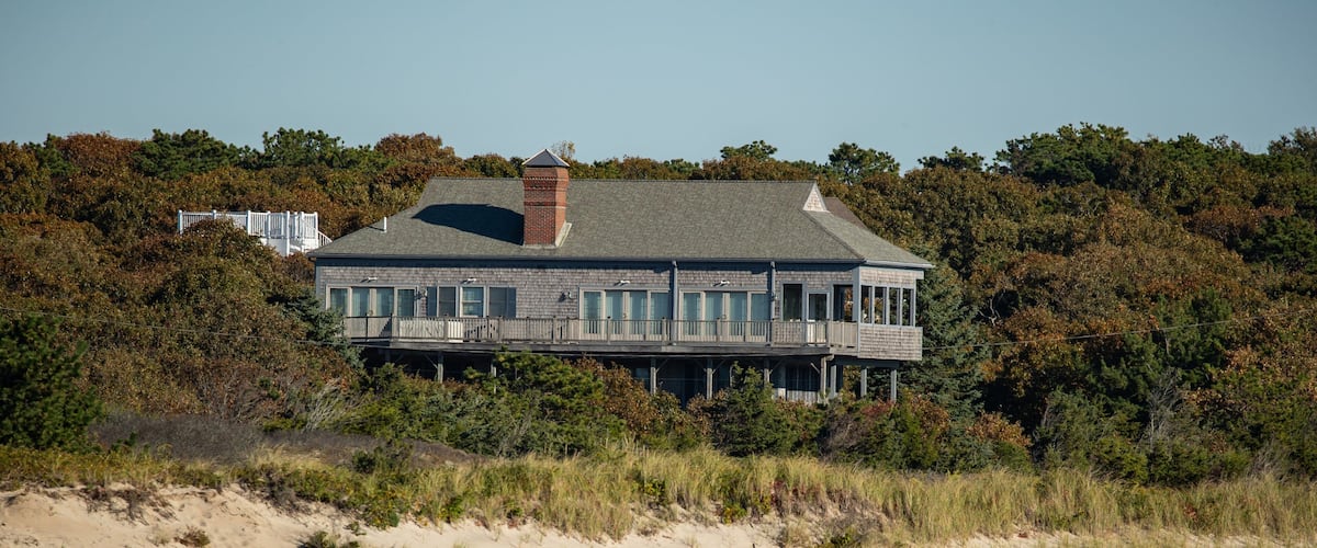 A coastal house with a wooden deck, surrounded by trees and a sandy beach.