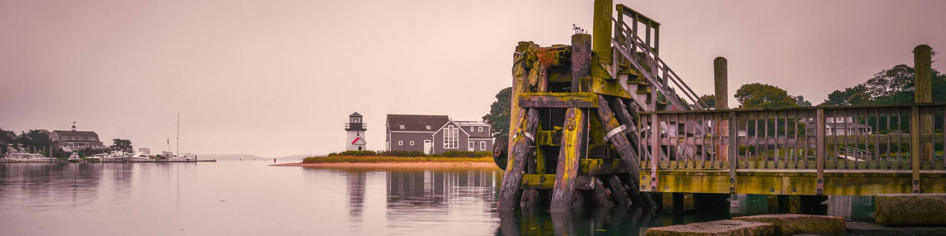 Pink sunrise seascape in Hyannis Harbor in Massachusetts. Lewis Bay Lighthouse, commercial dock, and stone bank with seawater reflections.