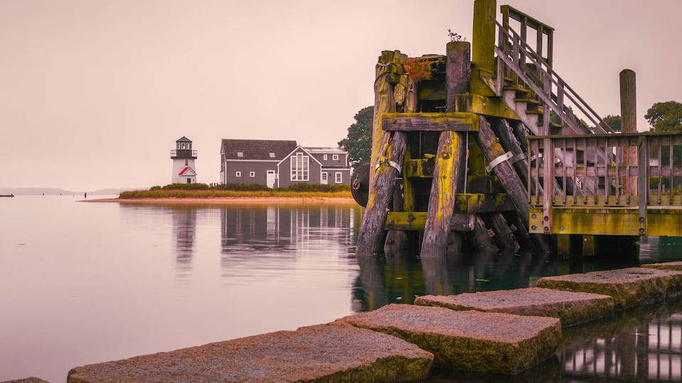 A calm waterfront with a lighthouse, a wooden structure, and a row of houses.