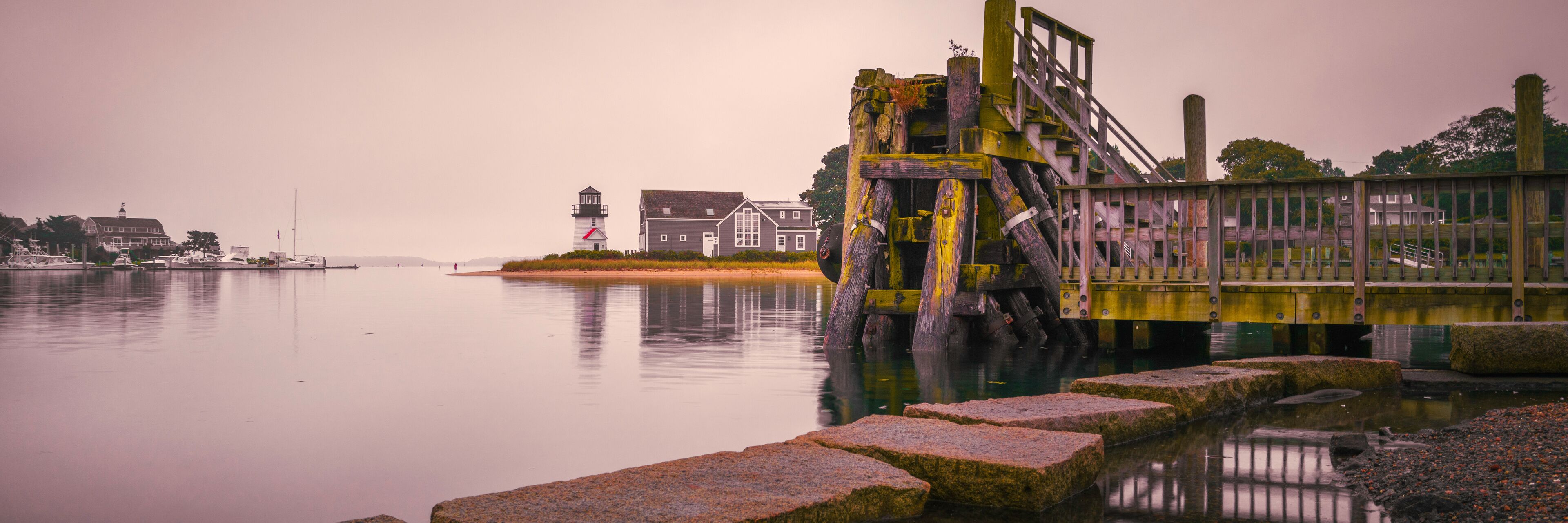 A calm waterfront with a lighthouse, a wooden structure, and a row of houses.
