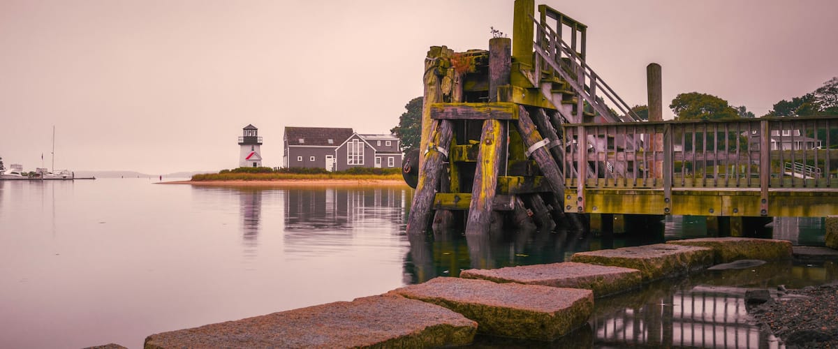A calm waterfront with a lighthouse, a wooden structure, and a row of houses.