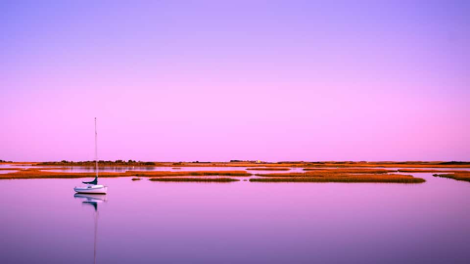 A calm water body with a sailboat anchored near the shore, surrounded by marshland and a clear sky.
