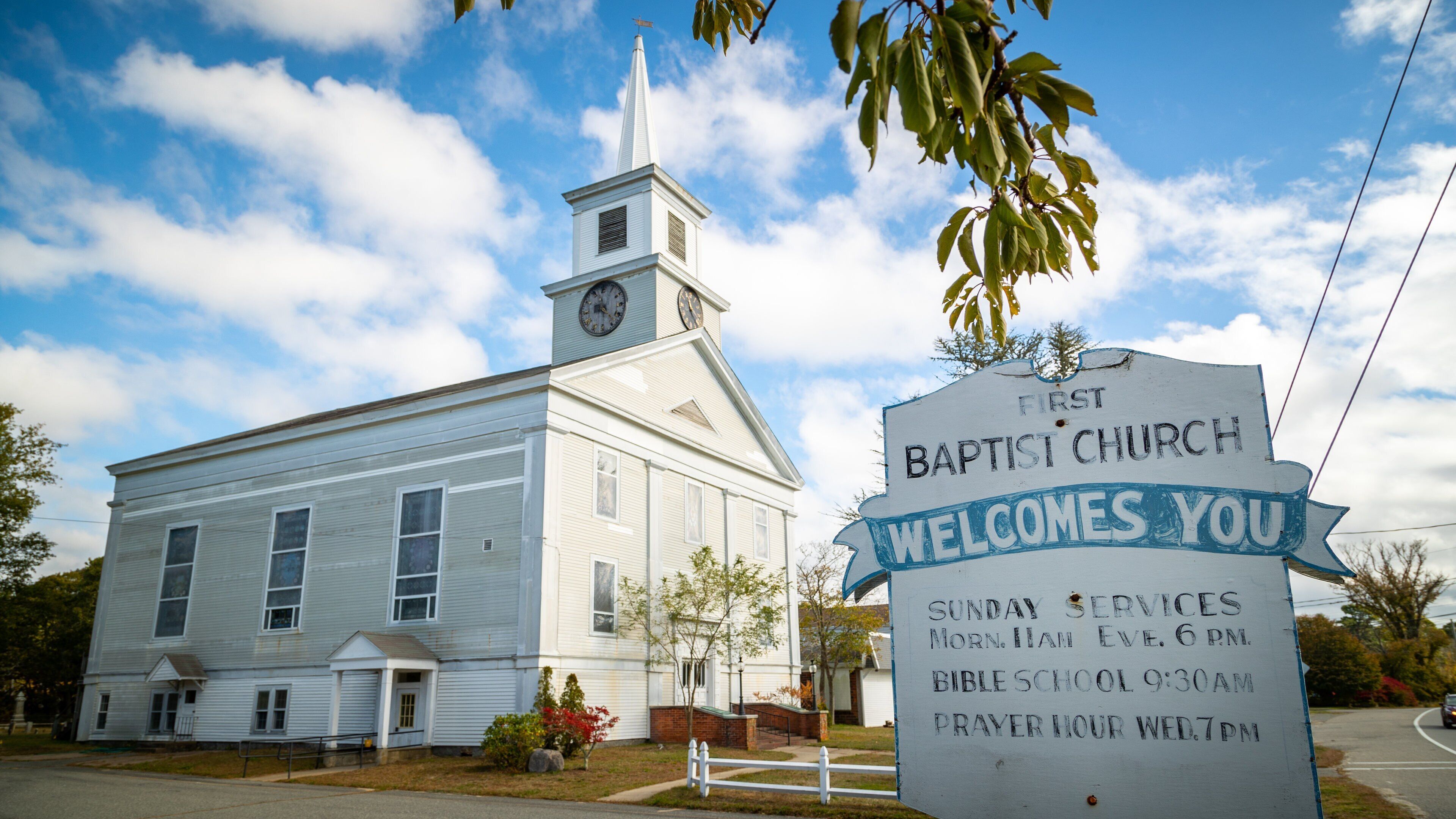 A white church with a steeple and a clock, a signboard with service times, and a clear blue sky.