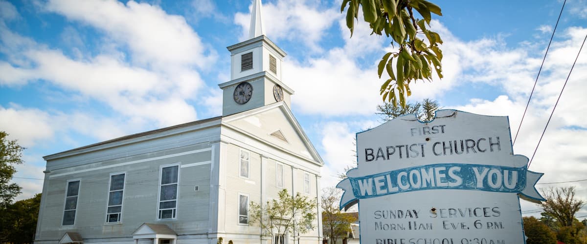 A white church with a steeple and a clock, a signboard with service times, and a clear blue sky.