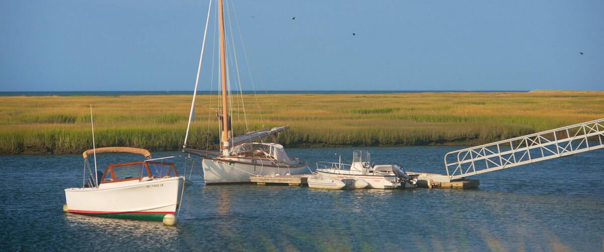 A calm waterway with several boats, including a sailboat and a motorboat, docked near a grassy marshland.