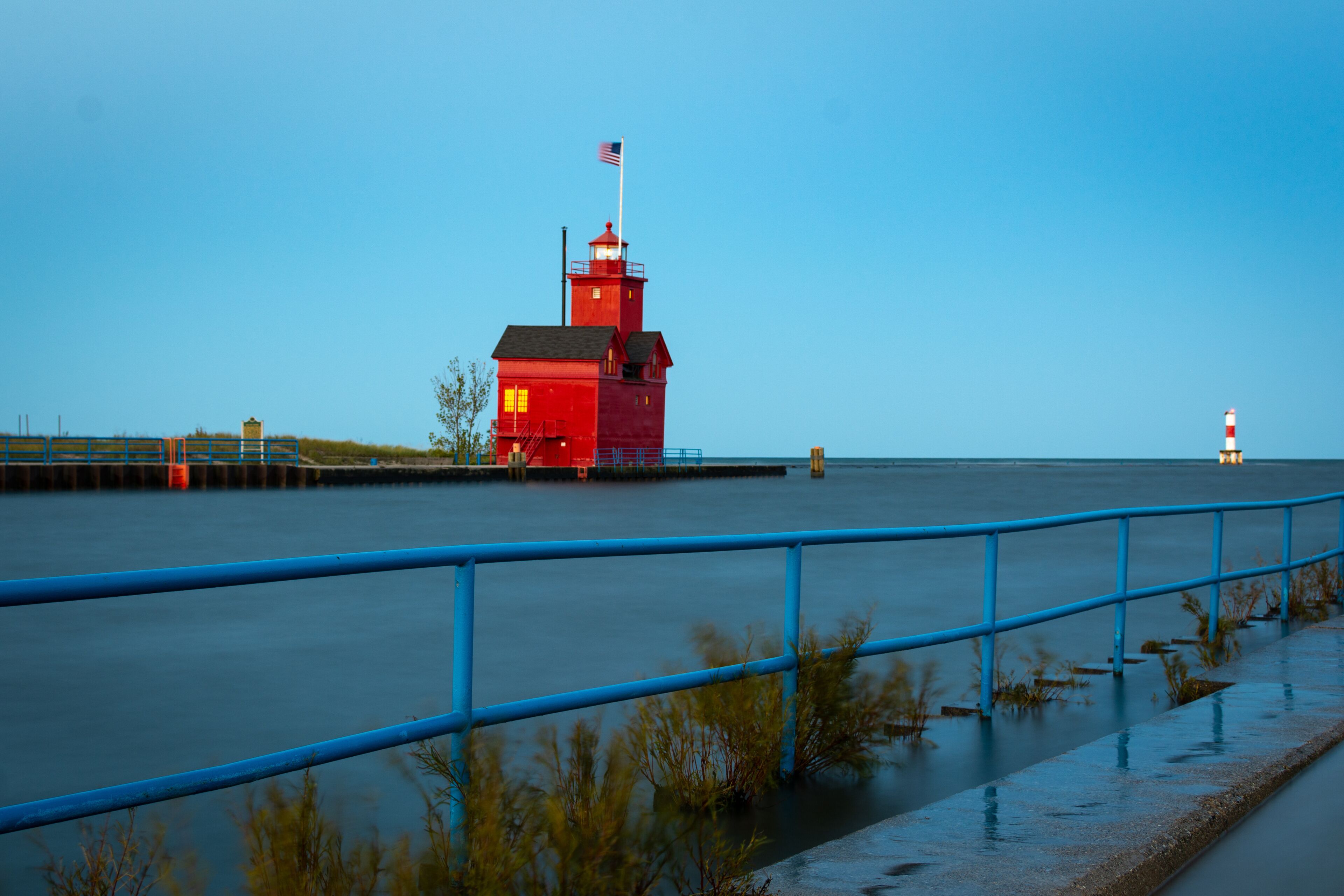 A red lighthouse with a flag, a blue railing, and calm water.