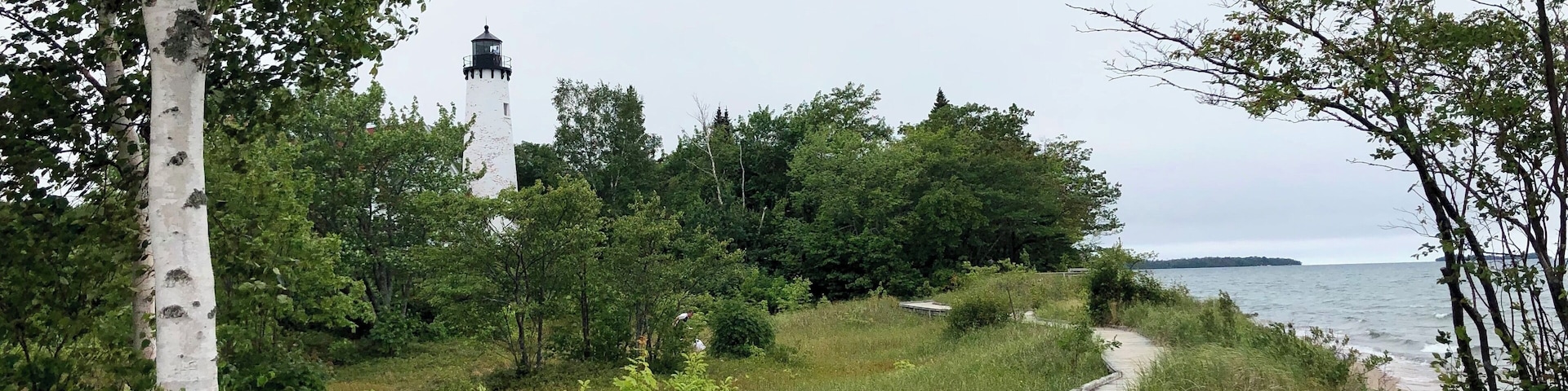 A wooden boardwalk leading to a lighthouse by the water, surrounded by trees and greenery.