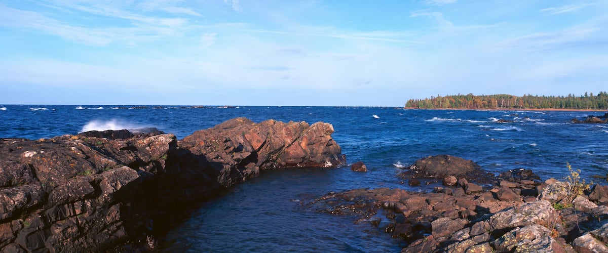 A rocky shoreline with waves crashing against the rocks and a forested island in the distance.