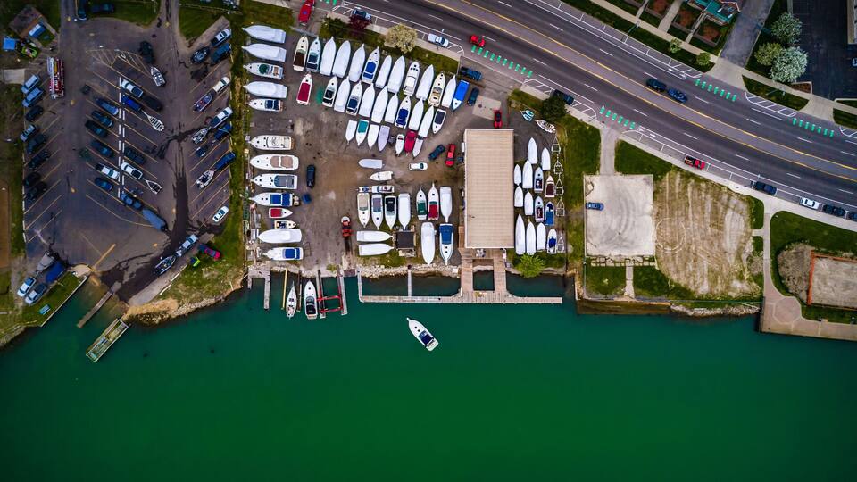 An aerial view of a marina with numerous boats docked, a parking area, and a road adjacent to the water.