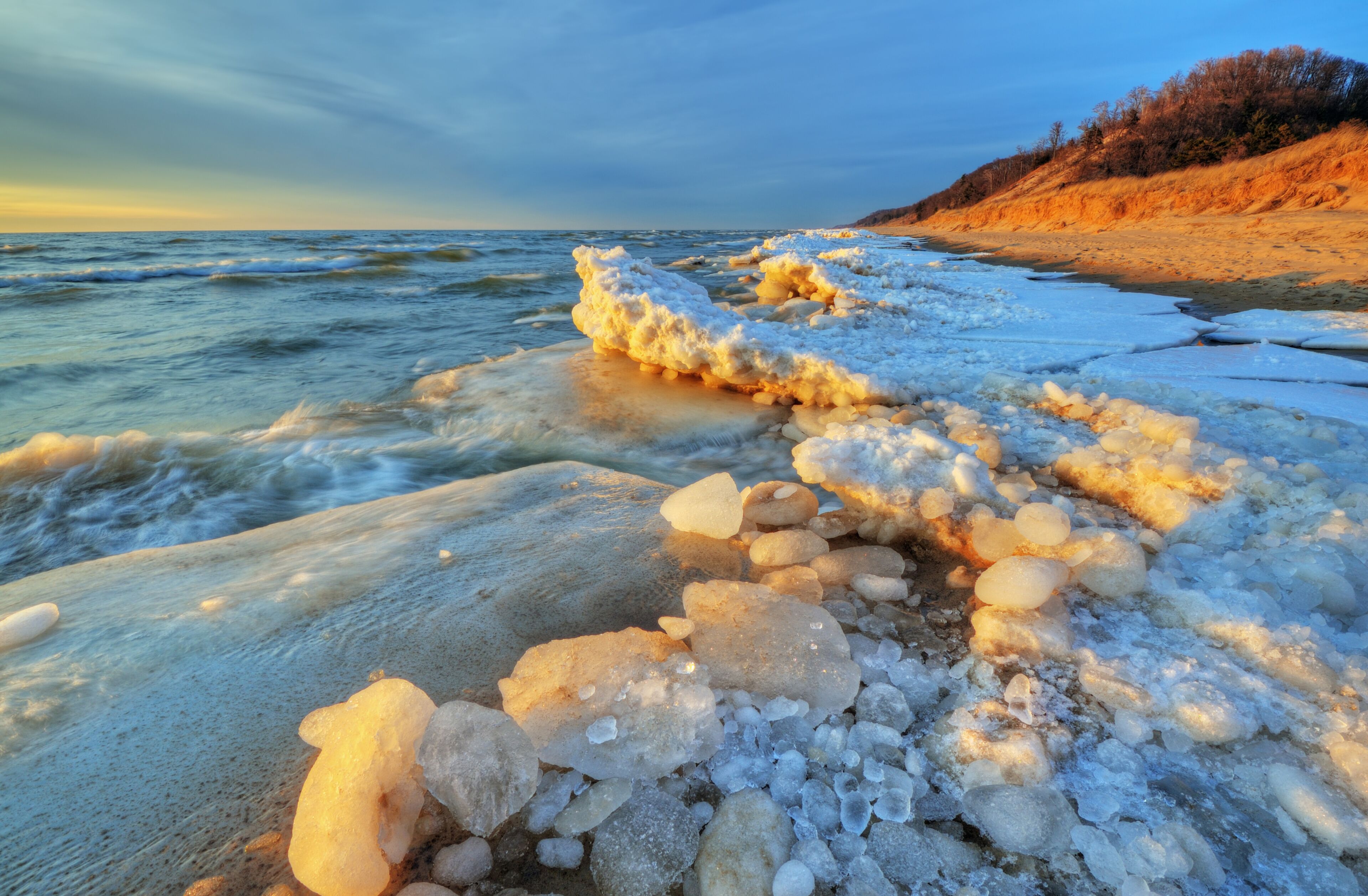 A frozen shoreline with ice formations and a distant forested hill.