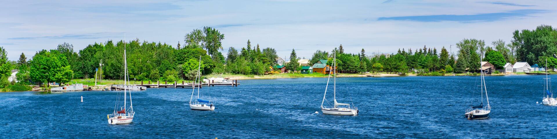 A calm lake with several sailboats anchored near the shore, surrounded by a forest and a small settlement.